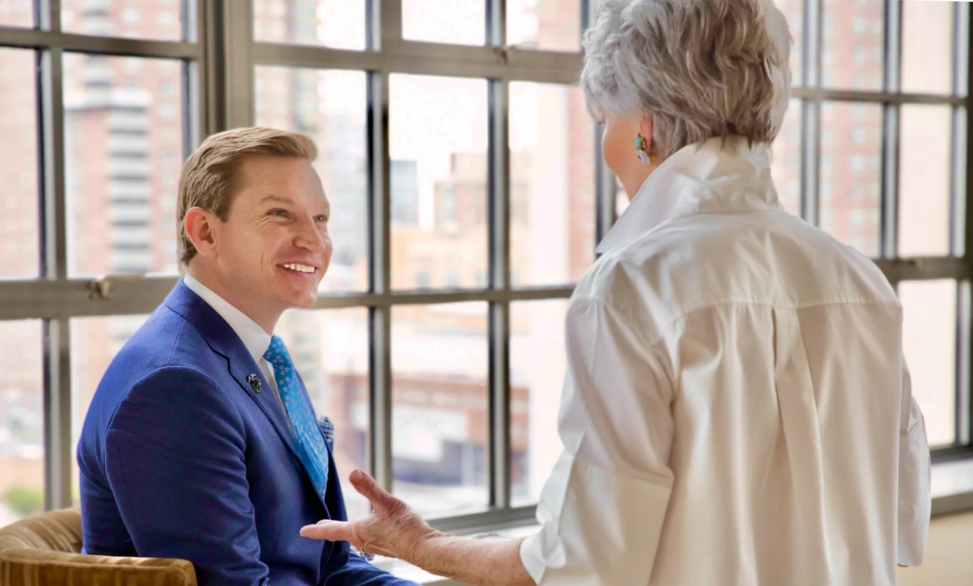 Brendan Hart seated in conversation with another person beside a wall of windows.